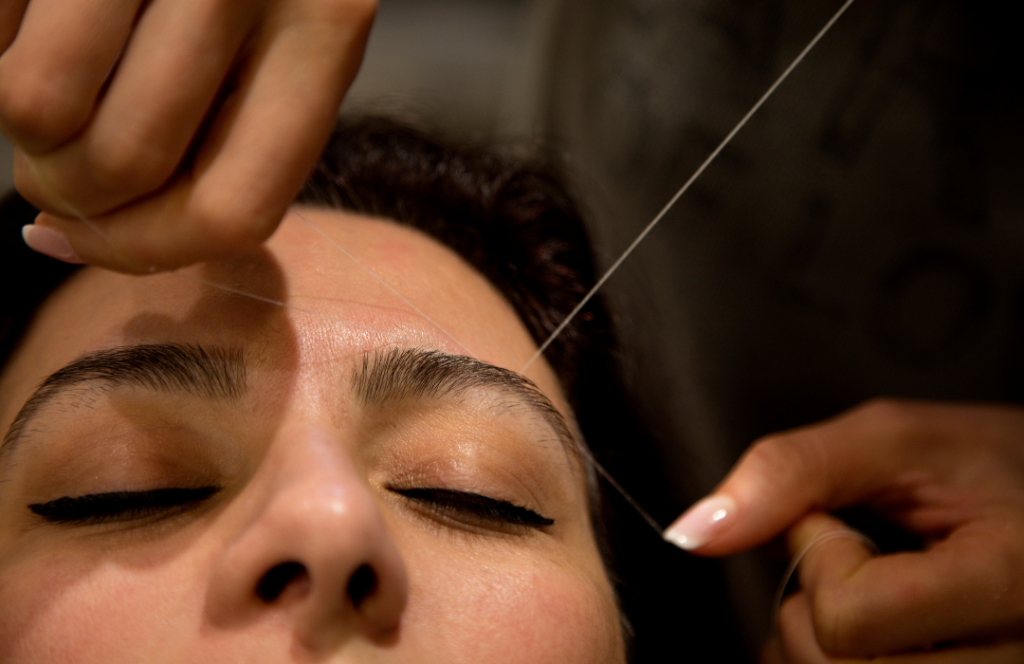 A woman sits in a salon chair as a beautician expertly threads her eyebrows, focusing on precision and detail.