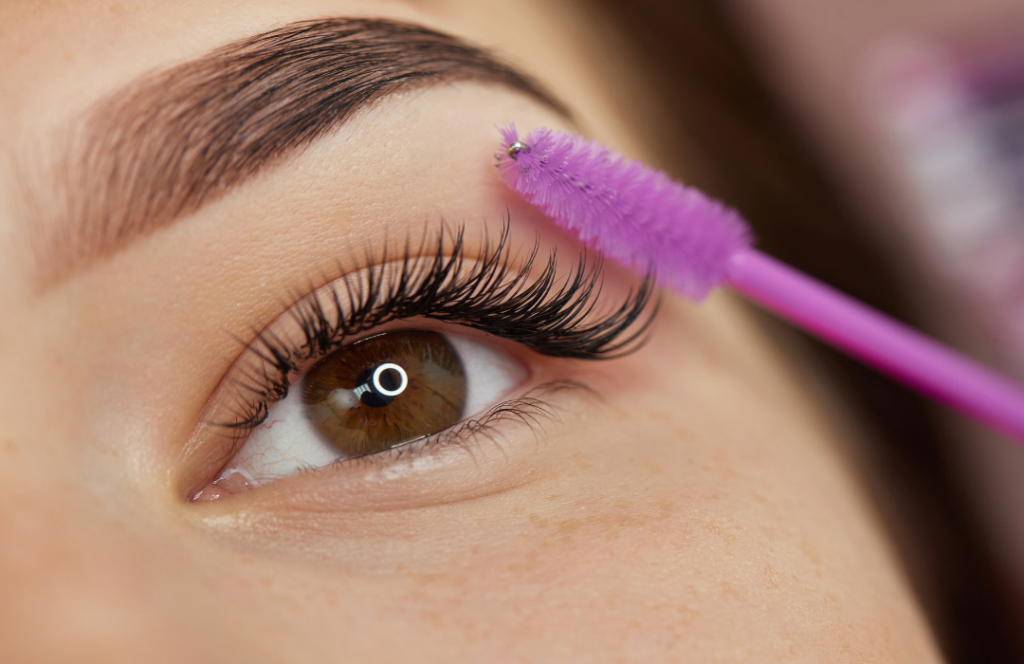 A woman with long eyelashes holds a pink brush, showcasing her beauty and elegance in a well-lit setting.